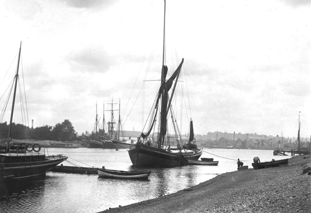 A photo of a river with a pebbley bank leading down to it. There are a number of sail boats moored in various places, including a large barge in the centre with its sails furled.