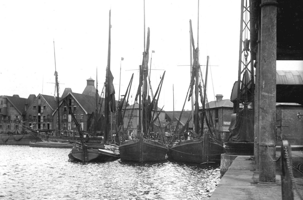A photo of the corner of some docks with five-storey warehouses lining the dock in the background. There are three barges docked next to each other, sails furled.