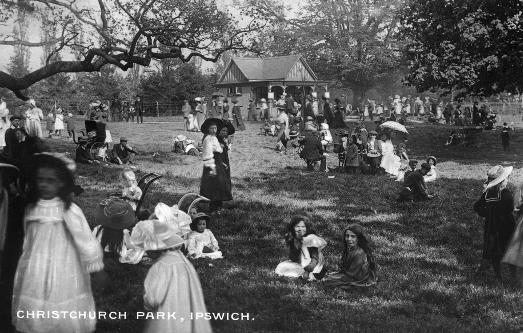 A photo of an area of grass and trees with a small single storey building in the background. The sun is casting shadows. There are lots of adults and children sitting, walking and playing. Women and girls in long dresses and, usually, wide-brimmed hats. The men most often in flat caps and suits.