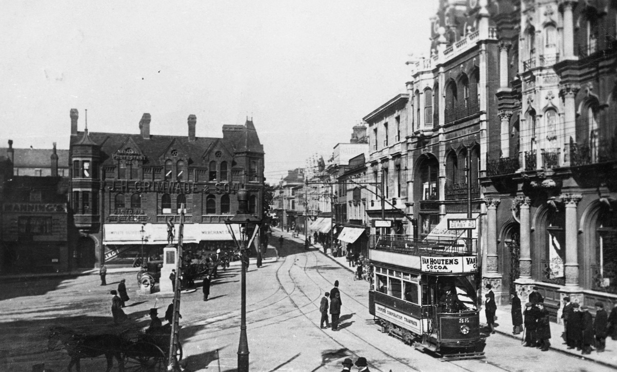 A photo of a wide junction in a town. The impressive stone buildings are three stories tall. Many have arched windows, pillars, and balconies. There is a double-decker tram and the only other vehicle is a two-wheel cart pulled by one horse. There are a few dozen people in dark clothes, mostly men.