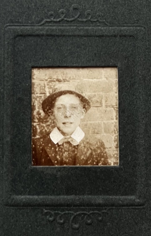 A photo of a bit of a photo album, black card with an embossed frame, and a square cut-out at the centre. Through the cut-out we see a sepia-toned photo of the head and shoulders of a young boy, possibly early teenage, facing the camera, in front of a brick wall. He's wearing a dark suit jacket, a dark shirt, with a prominent white colloar overlapping both the jacket and a bow tie. He's wearing a hat, that looks soft with a narrow brim. He's just about smiling.