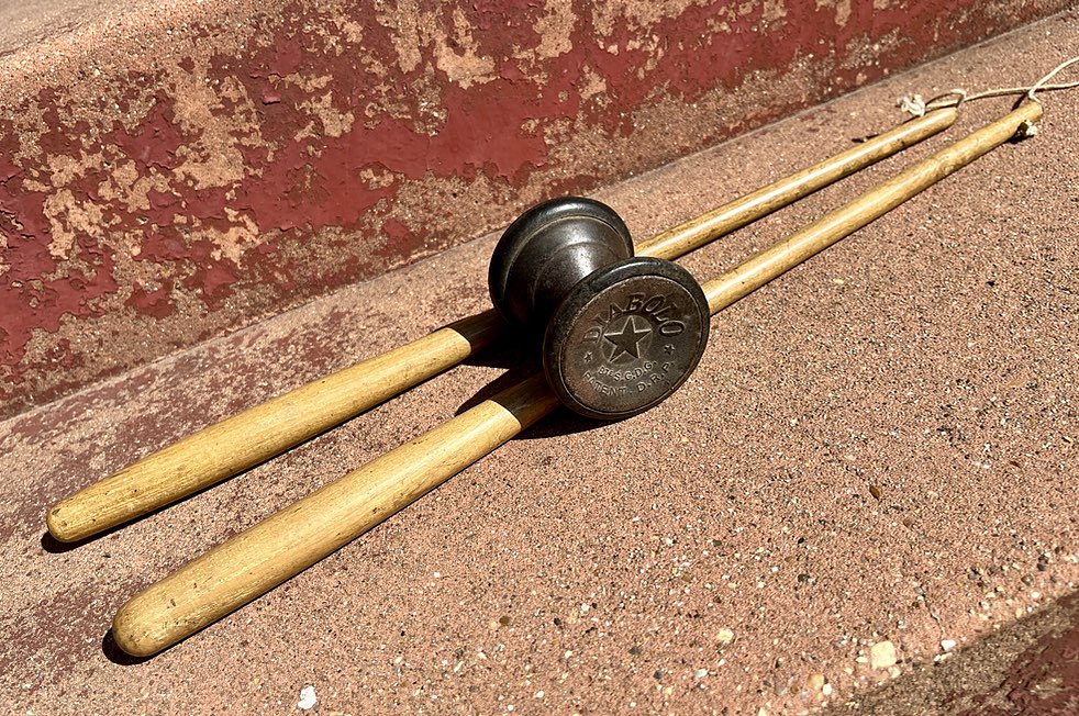 A photo of two round wooden sticks, tapering towards the far end where they have a piece of string tying them together, through holes in their ends. There's a dark metal thing, round, like two circular cones joined at their apexes. There is black rubber around the circular edges. The visible end has a five-pointed star in the centre and the word 'DIABOLO' embossed on it.