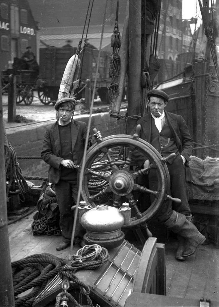 A photo of two men on an old barge, either side of a large wooden ship's wheel. They're both wearing flat caps, jackets and trousers, and each has a cigarette in his mouth. On the dockside behind them are carts and men.
