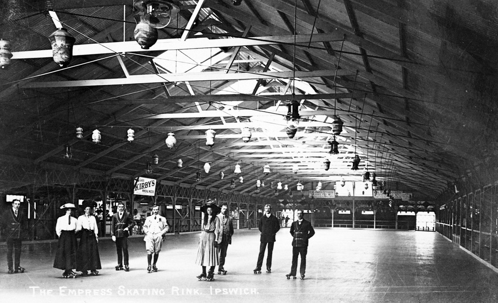 A photo of inside a large hangar-like building with a high pitched roof supported by many angled and horizontal beams. Nine adults stand on the flat empty floor facing the camera, all wearing roller skates. The men are wearing suits, three with what look like smoking jackets. The three women are in dresses or long skirts, with wide-brimmed hats. An advertisement in the background is for 'Kirby's Mineral Water'.