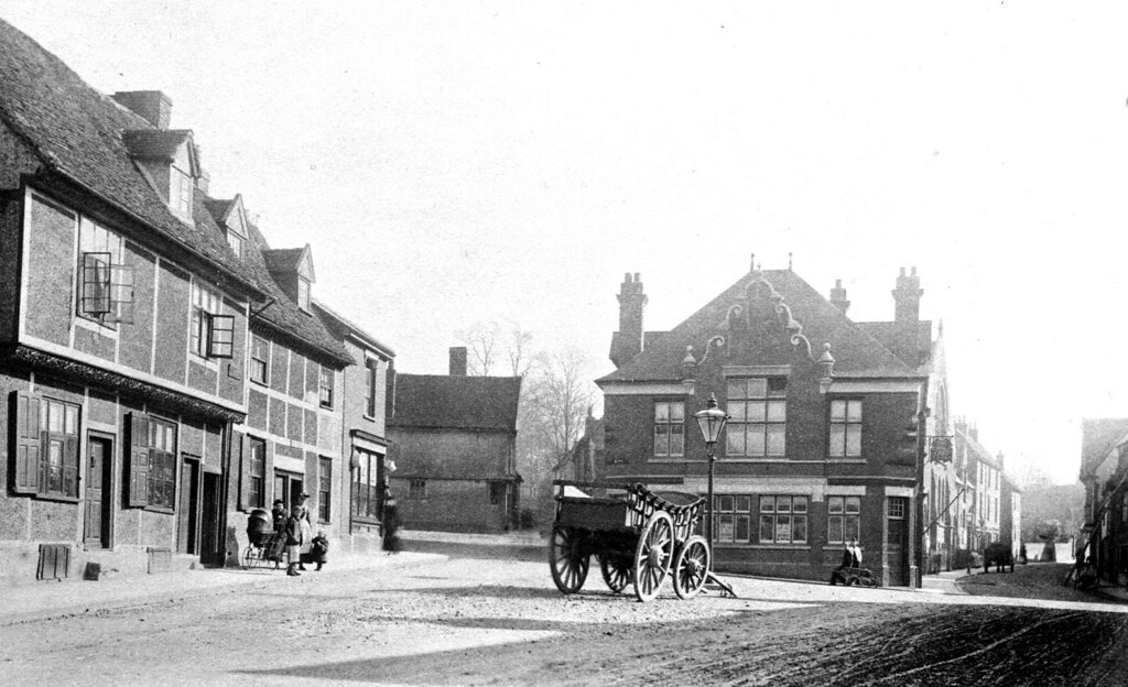 A photo of a wide junction on a street. There's a four-wheeled wagon with no horses stood in the open space. Behind it is a two-story building with a grand roof. There's no traffic on the roads and only a few people on the pavements.