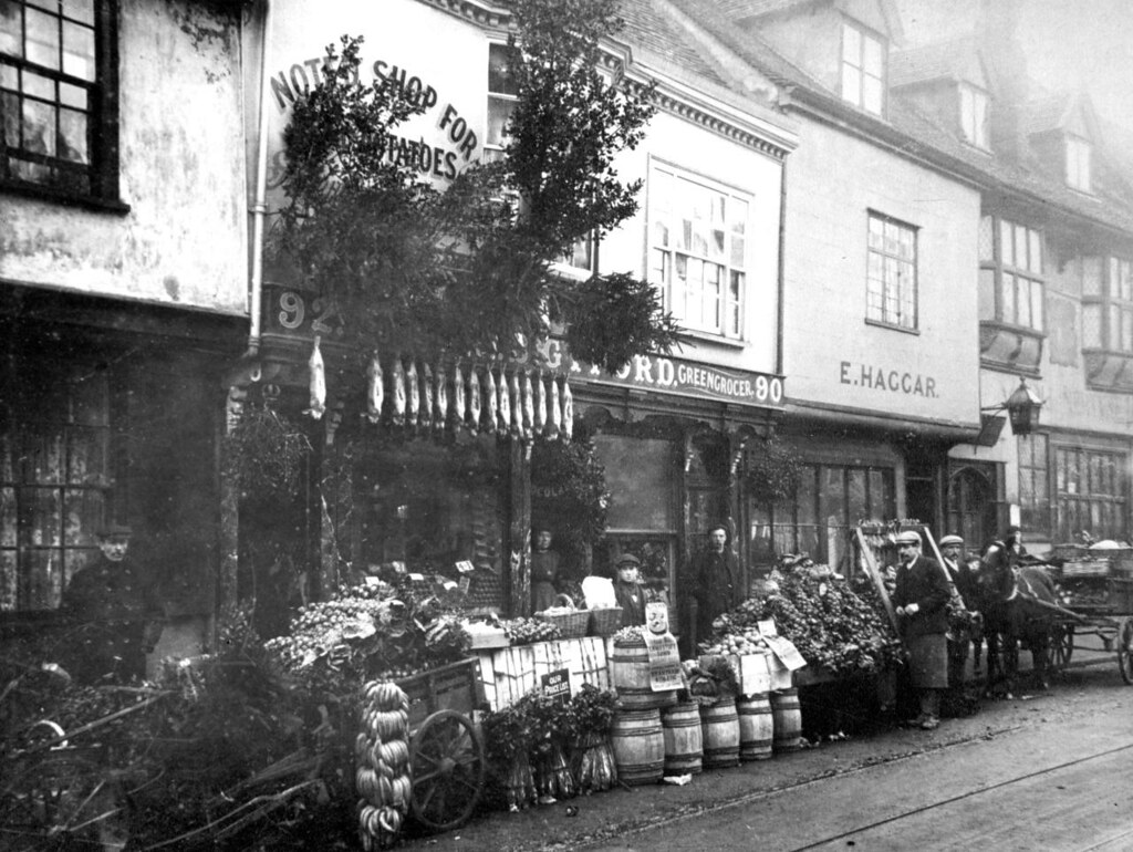 A photo of two shops on a street. 90-92 is C. B. Gyford, Greengrocer, its sign obscured by foliage presumably fixed to the shop. Outside are many barrels, a cart and displays of fruit and vegetables including bananas. Next door the shop has a sign reading 'E. Haggar'. Five men are standing around along with one boy and then a woman in Gyford's shop doorway.