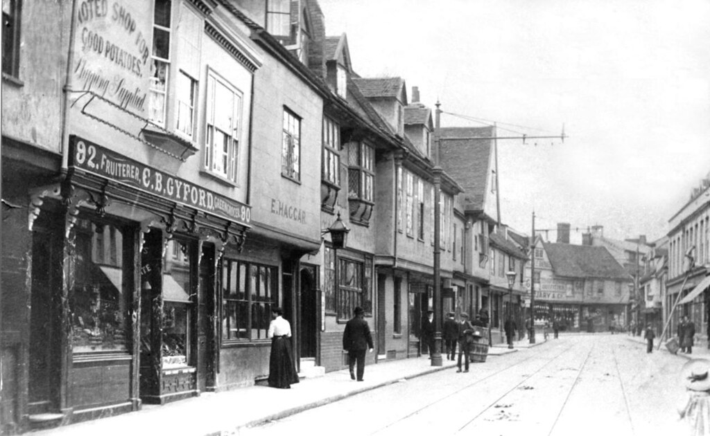 A photo of a street of old houses. The first floors mostly jut out slightly over the ground floors. Some have dormer windows in the roofs. The building closest on the left is numbers 90-92 and its sign reads 'Fruiterer. C. B. Gyford. Greengrocer. There are no vehicles but tram tracks are just visible on the road surface.