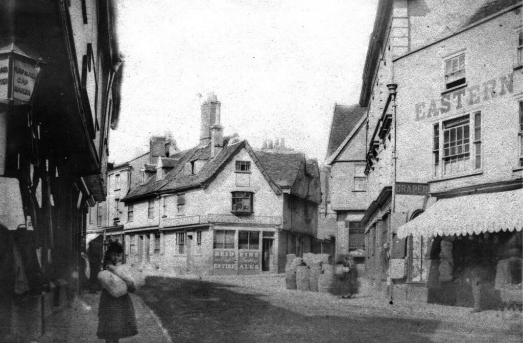 A photo of a junction on a street with old two and three storey buildings. There's no traffic and just a handful of people including a slightly blurry girl in a dress in the foreground carrying a large package.
