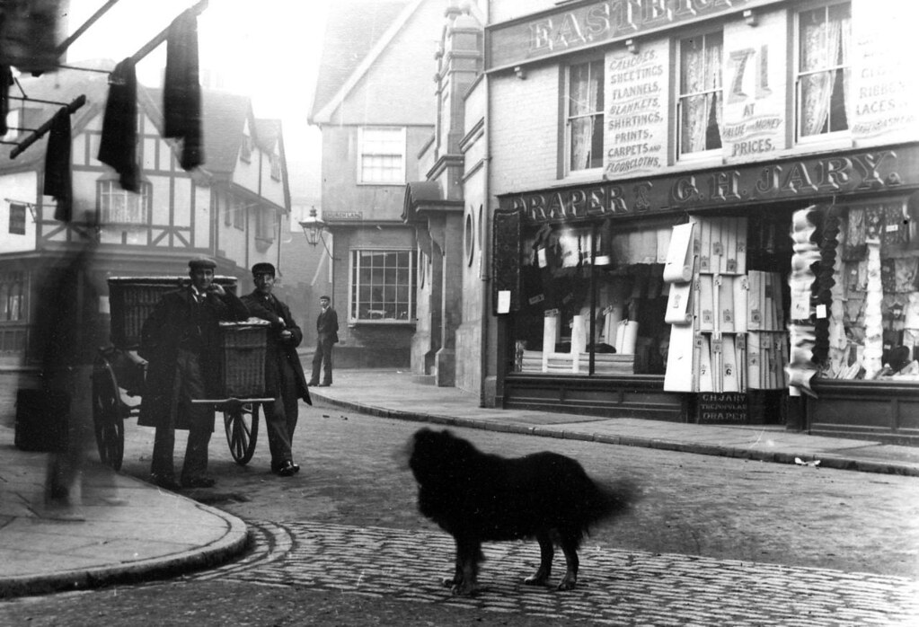 A photo of a cobbled street junction. There is a large, slightly blurred, dog in the foreground. Opposite the junction a shop has the sign 'Draper. G. H. Jary'. Four men are standing around, wearing caps, one of them very blurred. One of the buildings in the background is half-timbered.