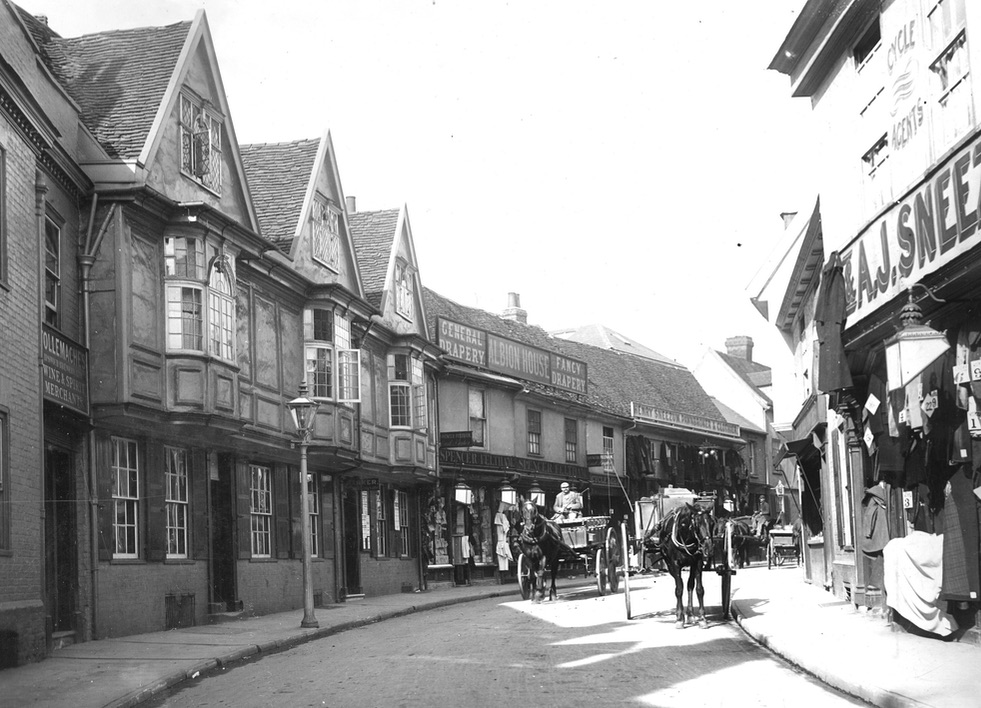 A photo of a street of two and three storey buildings. On the right we can see part of a shop and its sign 'A. J. Snee&hellip;'. In the distance is 'Henry Sneezum, Pawnbroker and Clothier'. Closer than that is 'Albion House. General Drapery. Fancy Drapery.' Between them is 'Cycle Agent'. There are two horse-drawn carts on the road.