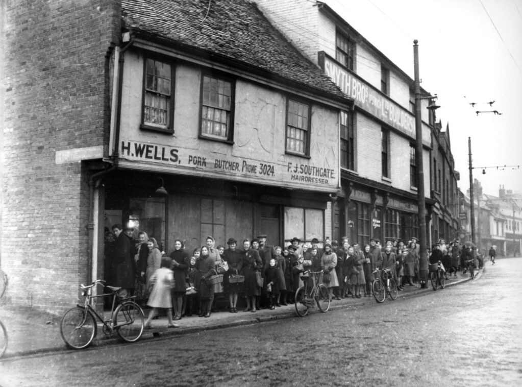 A photo of one side of a street of old two and three storey buildings. The closest is H. Wells, a 'pork butcher' with a huge queue of people leading from the open door away down the street.