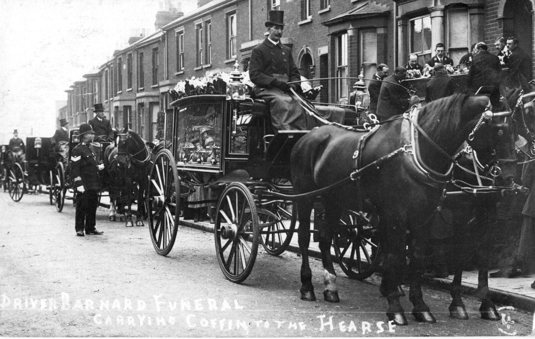 A photo of a street of terraced houses with three or four horse-drawn carriages driven by men in overcoats and top hats. The front-most one has two dark horses and is a hearse, with glass sides and flowers on top. In the background nine men carry a coffin down steps from the front door of one of the houses. A police sergeant looks on.