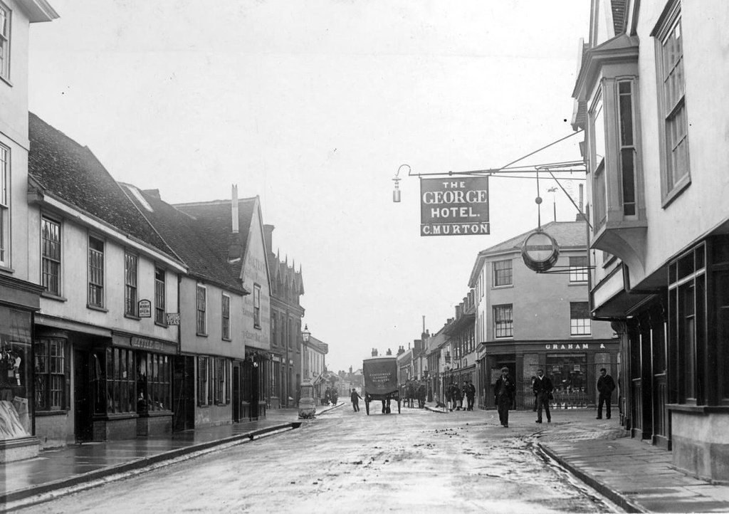 A photo of a wet town street with two and three storey buildings on each side. There are two horse-drawn wagons in the distance and a few people dotted around. A sign sticking out from the building closest on the right reads 'THE GEORGE HOTEL. C. MURTON'.