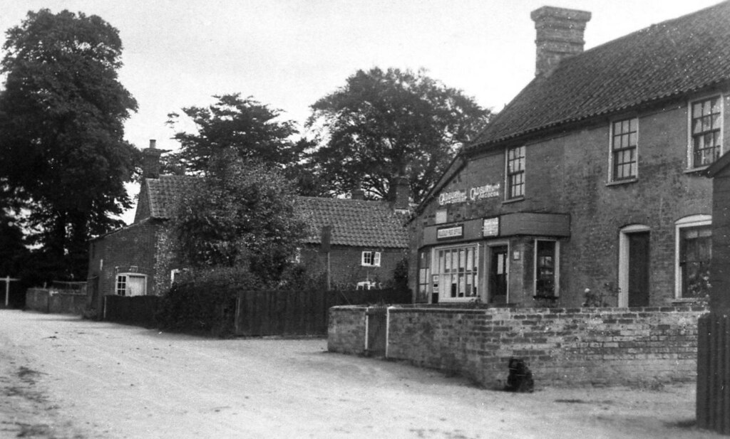 A photo showing a village road with a couple of brick buildings, and tall trees in the background.