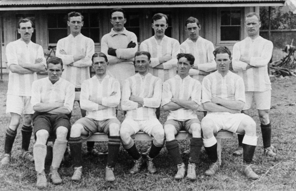 A photo of eleven men in two rows, the front row of five sat on a wooden bench. They're all wearing probably white tops with a wide, pale vertical stripe. Their shorts and socks are a variety of shades.