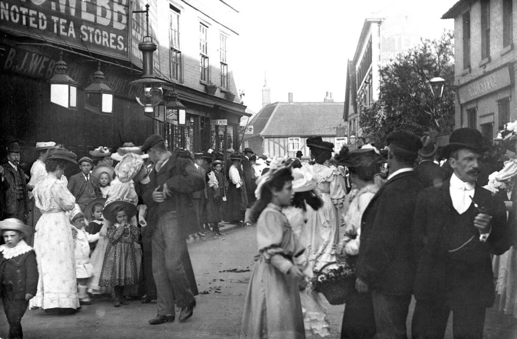 A busy street scene with a few dozen people quite dressed up. Women and girls are wearing long dresses and wide-brimmed hats. Men are wearing a variety of caps, boaters, bowler hats, etc. A man in the foreground with a bowler hat has a white shirt and tie, a three-piece suit, and is smoking a pipe. A small boy is looking at the camera, wearing a wide straw hat with a white collar spreading over his shoulders and chest.
