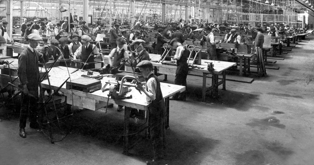A photo of a vast warehouse type room filled with many rows of workbenches. There are many men and boys at work on bits of metal. They're all wearing flat caps and either jackets or waistcoats.