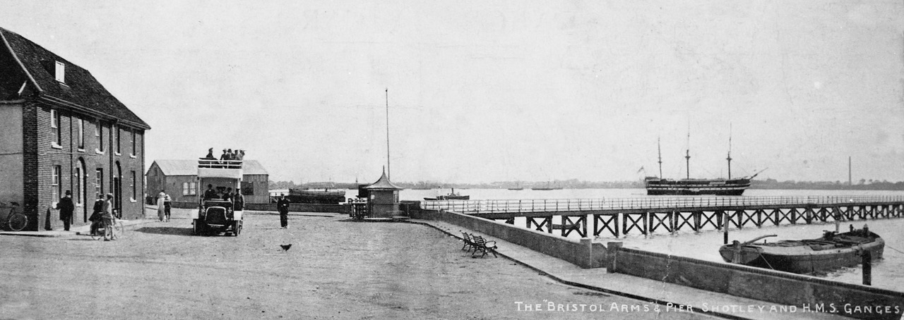 A panoramic postcard photo of the waterfront. On the left are two two-storey brick buildings. There's a wide almost empty street with one double-decker, open-topped motor bus, and a few people. There's a long narrow, empty pier stretching out into the water. In the distance is an old three-masted sailing ship.