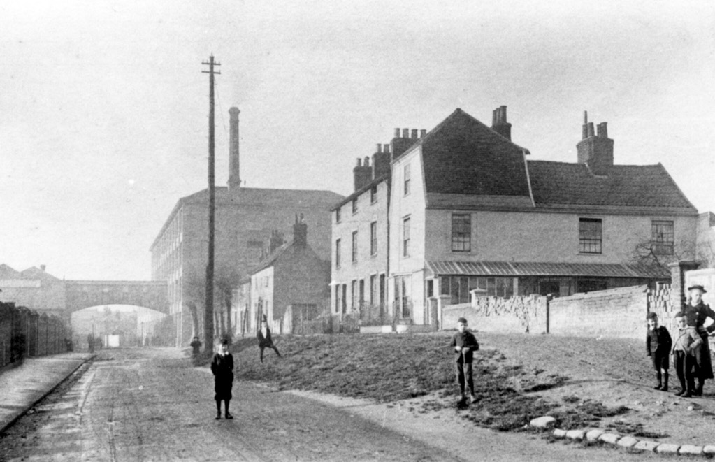 A photo showing an empty street. On the right of the road a grass verge leads up to the pavement and a few houses. In the distance is a four-storey warehouse or factory with a tall chimeny and a covered bridge across the road. There is a very tall telegraph pole. There are six young boys and a young woman stood at various places looking at the camera.