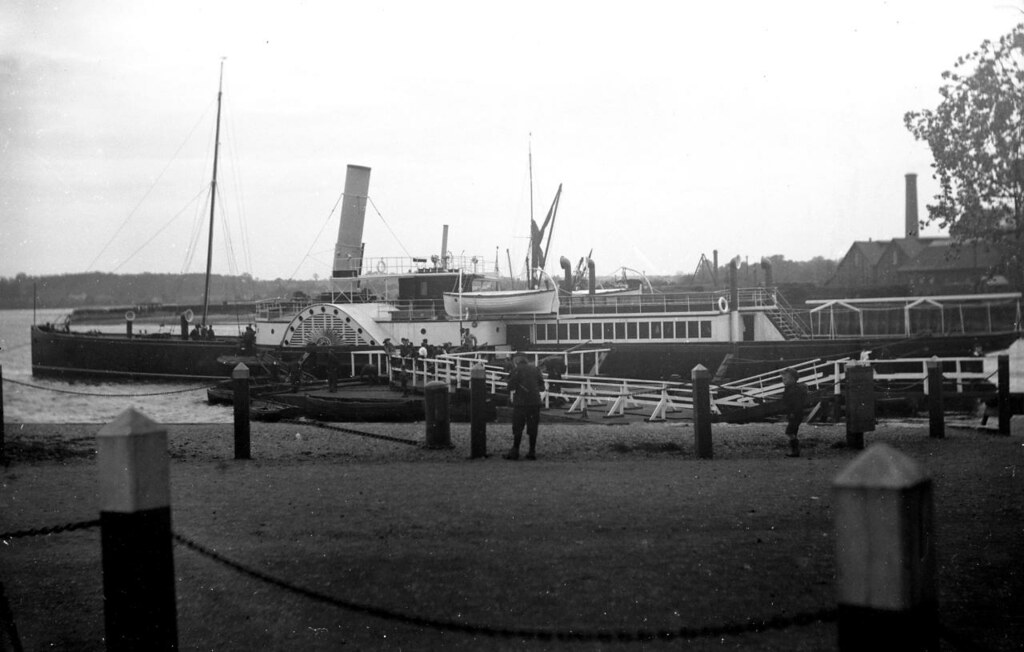 A photo looking across grass at a stretch of water. Moored at a jetty is a long, low ship with a tall, straight funnel. A semi-circular shape indicates the position of a paddle wheel.