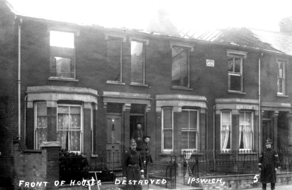 A photo showing a terrace of houses with no roofs and daylight showing through the upstairs windows. Policemen stand outside.