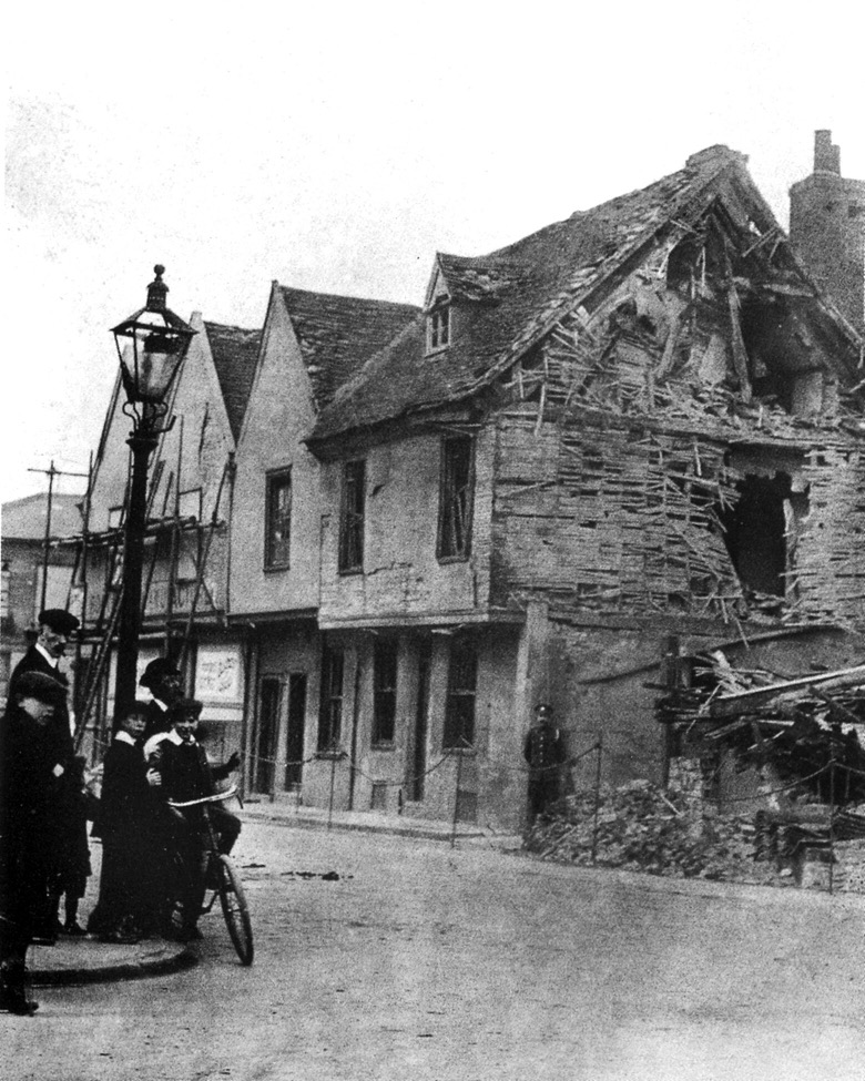 A terrace of houses with the end demolished exposing a large hole and wooden lath in the remaining building. A man in uniform stands guard and two men and three boys look on from the opposite side of th